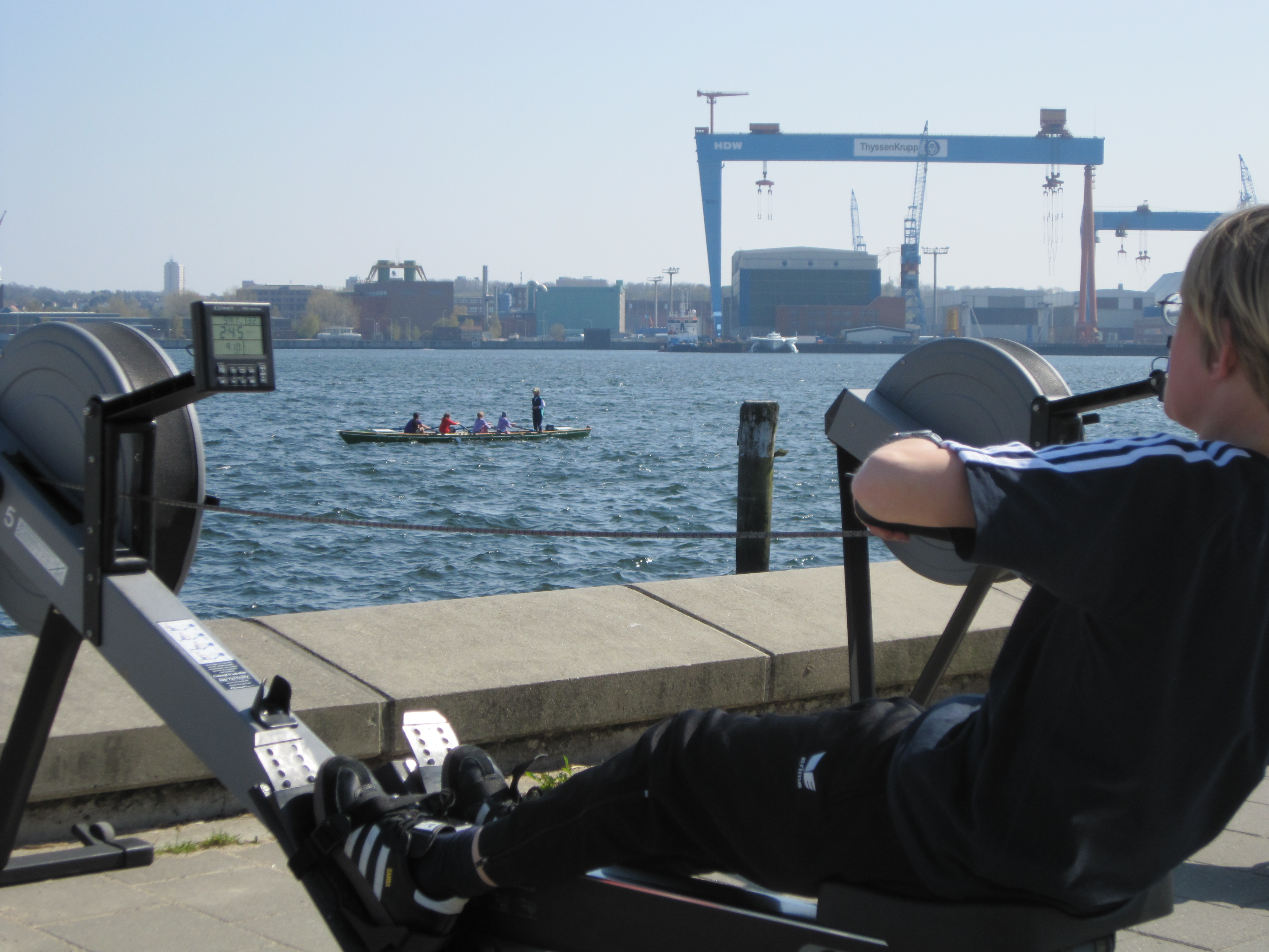 Kind auf dem Ergometer, im Hintergrund Kinder im Ruderboot vor dem Portalkran von German Naval Yards